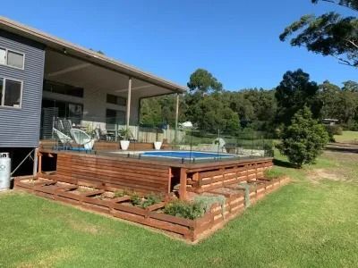 Wooden deck with above-ground pool and low garden beds in front of a house on a sunny day.