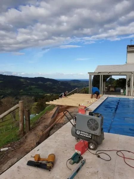 Construction of a wooden deck near a pool, overlooking a scenic valley under a partly cloudy sky.