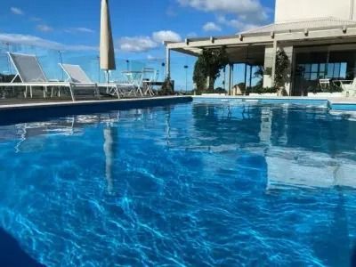 Swimming pool with blue water reflecting the sky, lounge chairs, and a building with a pergola.