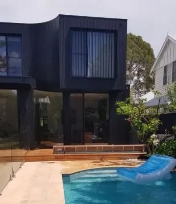 Modern black house with pool; blue float in pool, wooden deck, glass railings, and white house in background.