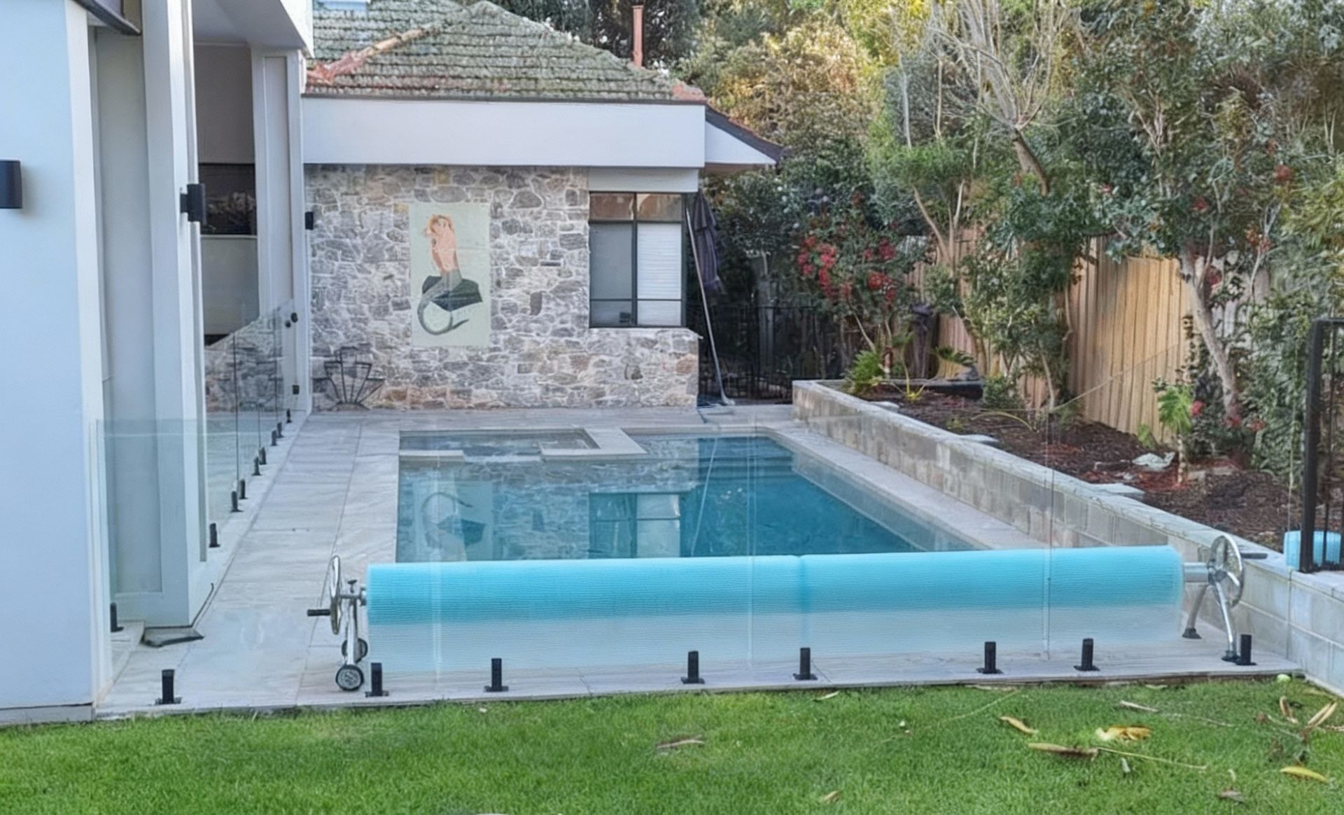 Swimming pool with blue cover, stone and glass fencing, near a house with greenery.