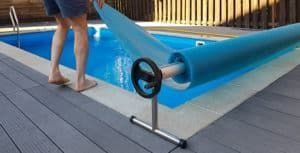 Person rolling a blue pool cover off a rectangular pool on a wooden deck.