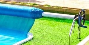 Blue pool cover rolled up on a metal reel, resting on green turf.
