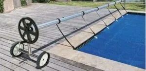 Pool cover being rolled up on the deck. Blue pool water. Silver and black cover roller.
