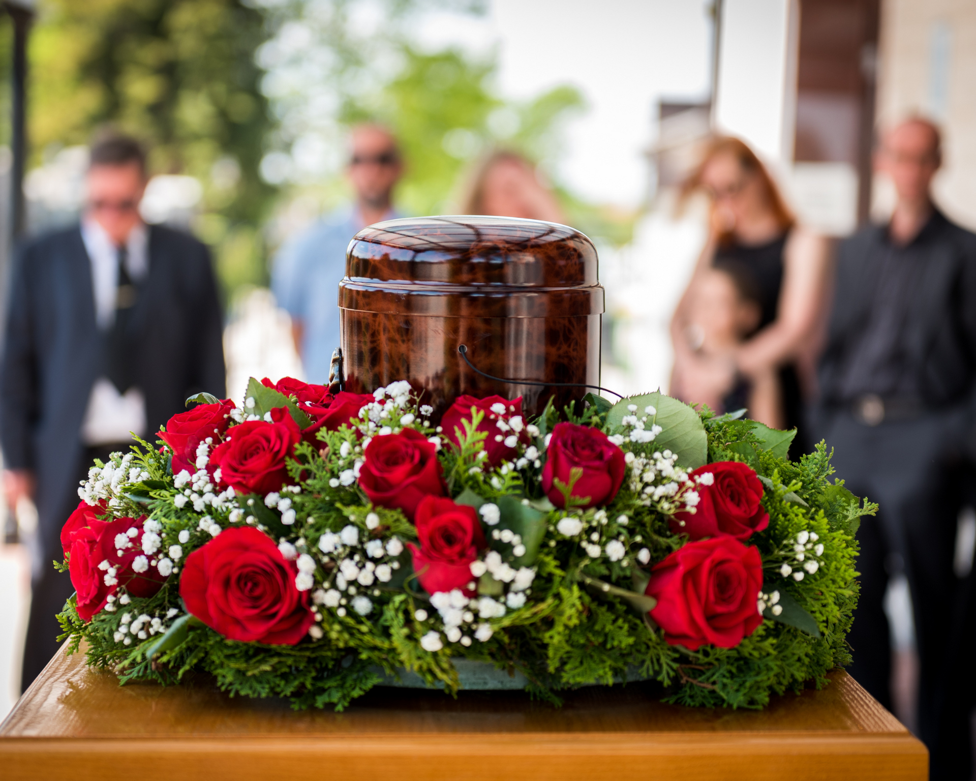 A dark wooden urn sits atop a bed of red roses and greenery, with blurred figures in the background at a funeral.