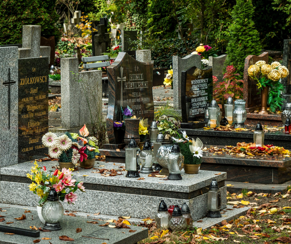 A collection of granite headstones adorned with fresh flowers and votive candles in a cemetery filled with autumn leaves.