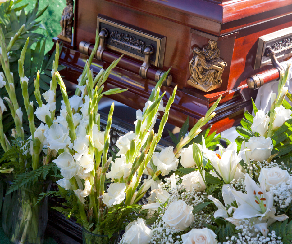 A dark wooden casket decorated with a gold figure, surrounded by white gladioli and roses.