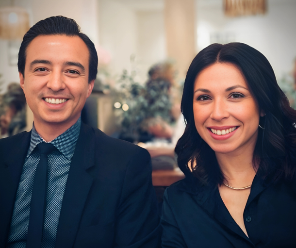 A man in a suit and tie and a woman in a dark shirt sit side-by-side smiling in a softly lit, casual dining setting.