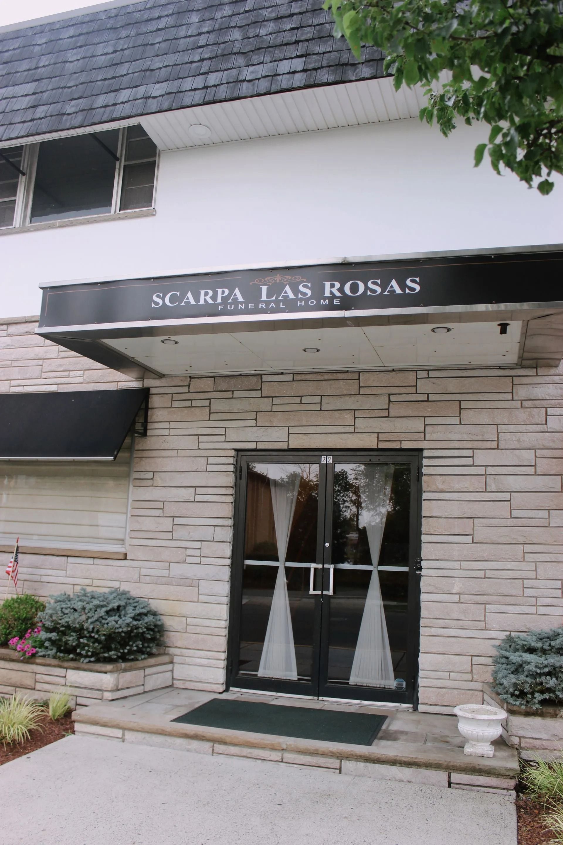 The exterior entrance of Scarpa Las Rosas funeral home, featuring a black awning over glass doors and light stone siding.