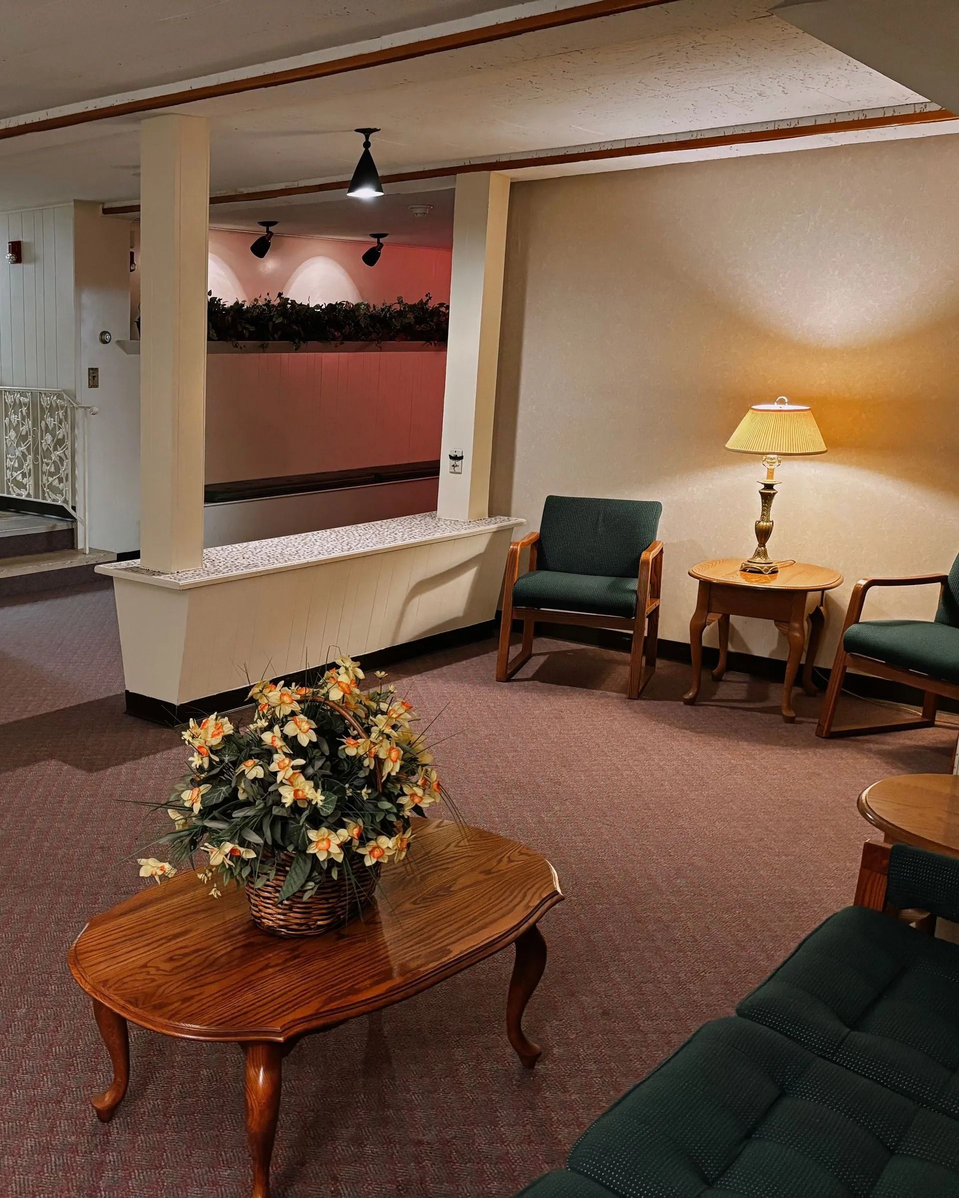 A waiting room with green armchairs, a wooden coffee table with flowers, and a lamp on a side table.