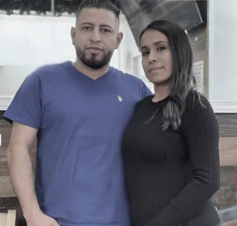 A man in a blue t-shirt stands beside a woman in a black long-sleeved shirt, both posing together against a wood wall.