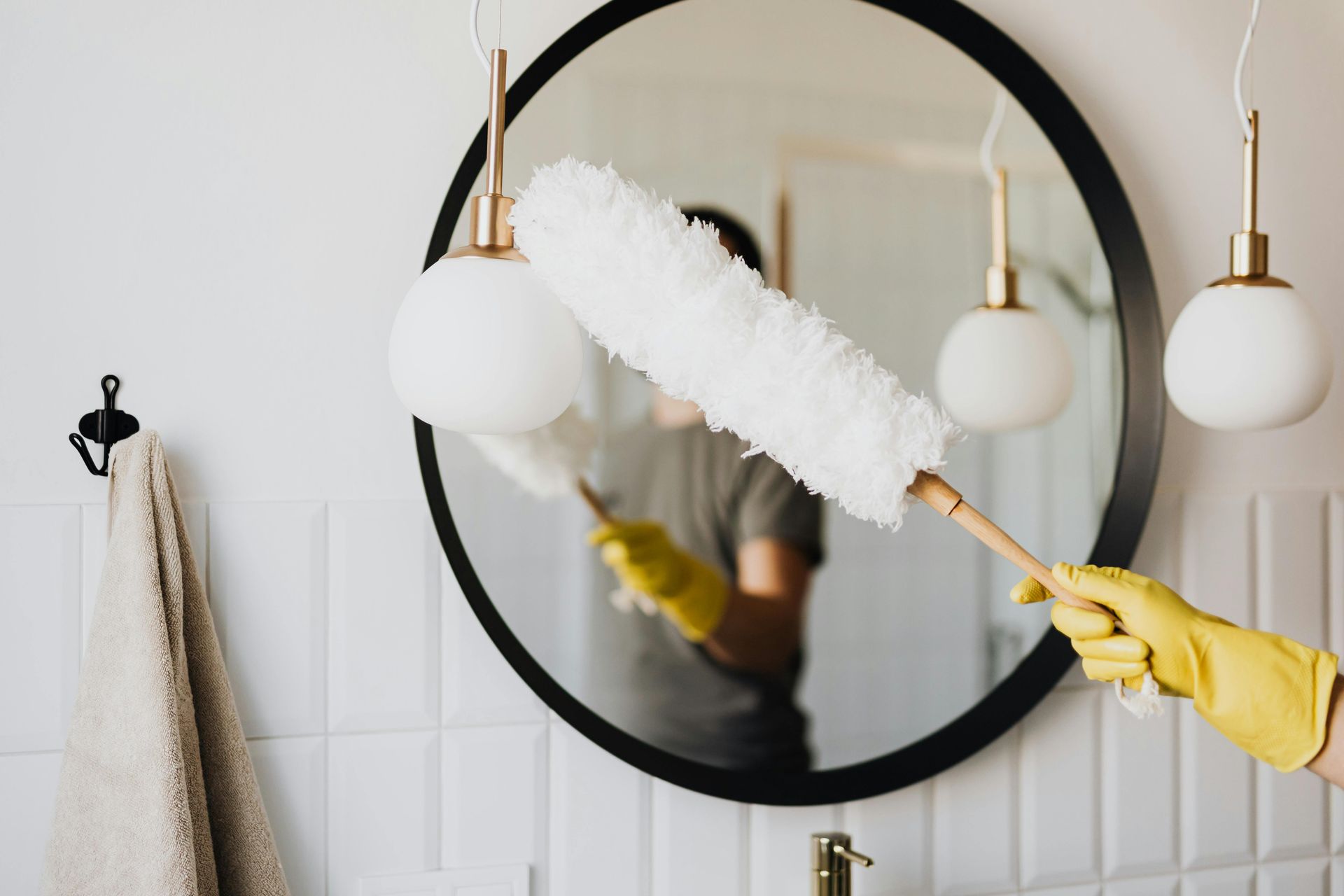 Person wearing yellow gloves dusting a circular mirror with a fluffy duster in a bathroom.