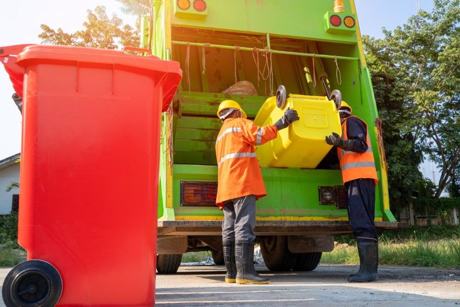 Garbage truck workers emptying a yellow bin beside a red trash can outdoors
