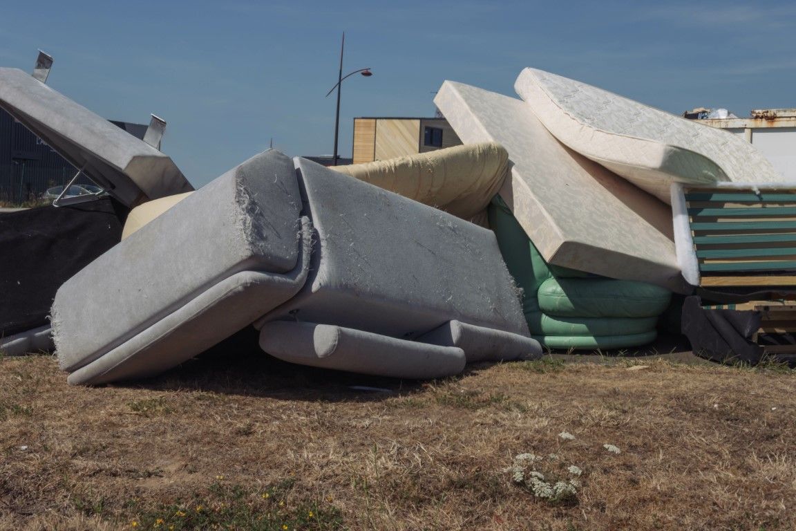 Discarded mattresses piled in an outdoor lot on dry ground, with a building and debris behind them.
