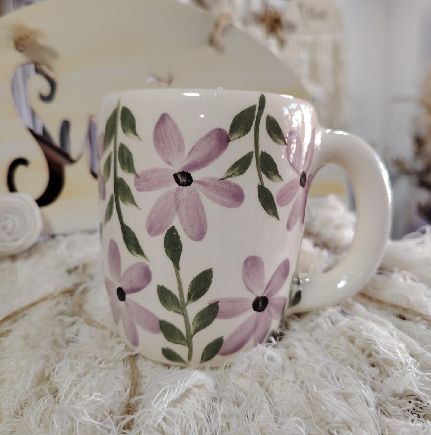 Taza de cerámica blanca con flores moradas y hojas verdes pintadas a mano, colocada sobre tela de encaje.