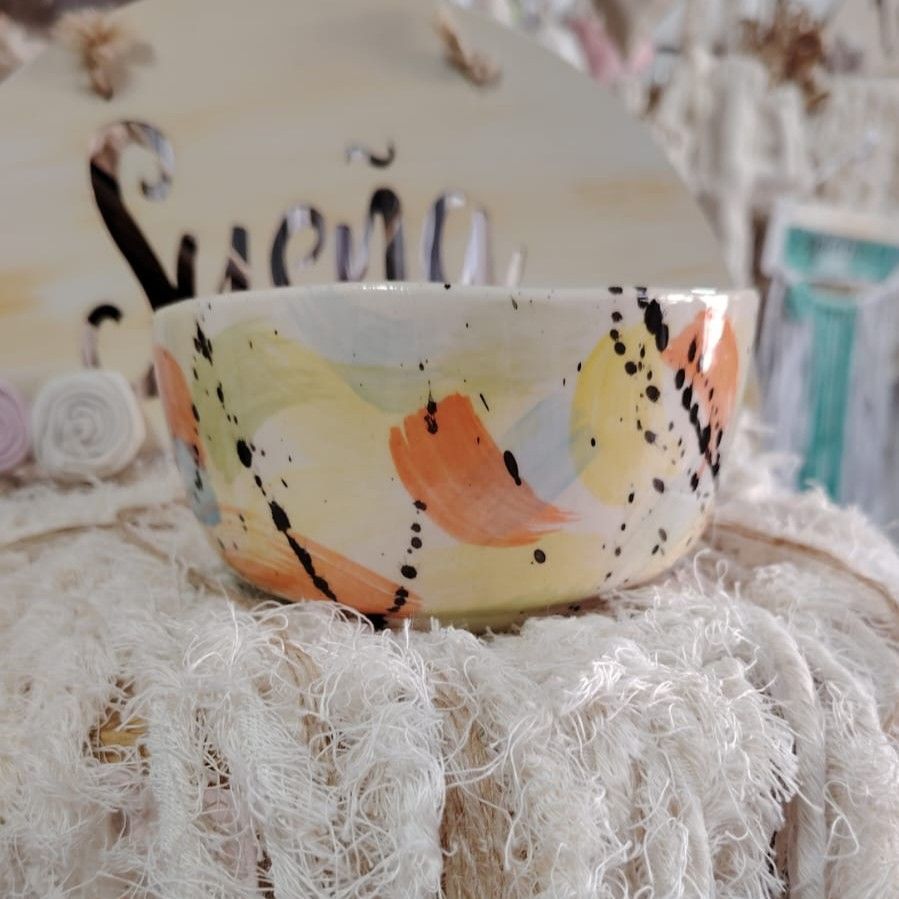 Cuenco de cerámica con pinceladas abstractas de colores sobre un paño blanco con volantes, delante de un cartel de madera.