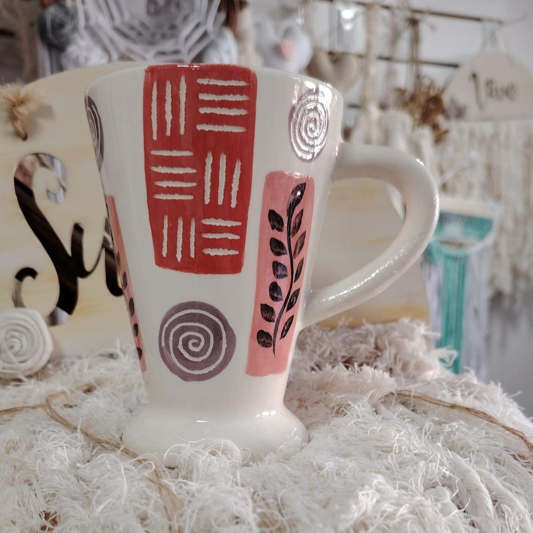 Taza de cerámica blanca con diseños estampados en rojo, rosa y gris, sobre una tela tejida a crochet de color blanco.