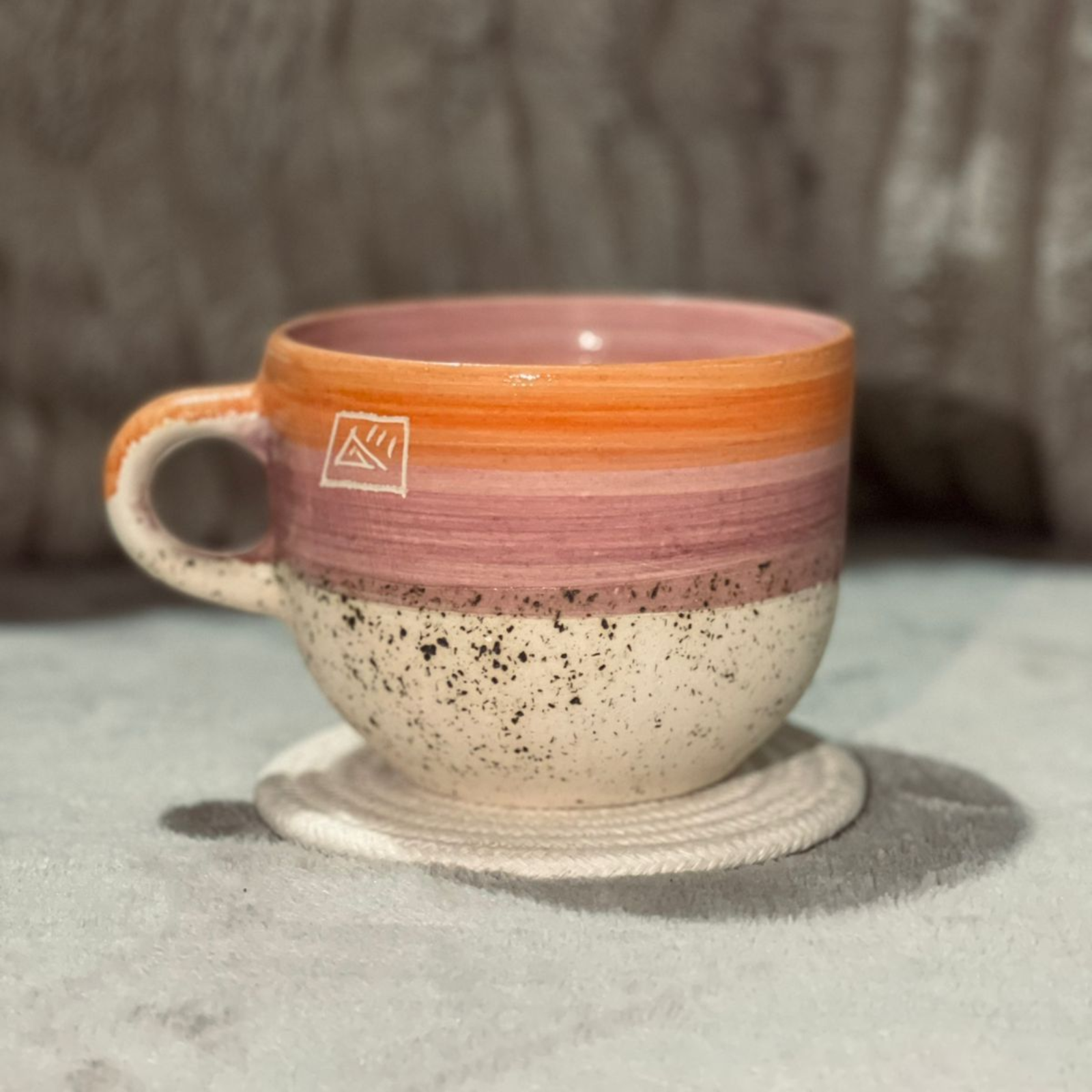 Una colorida taza de cerámica sobre un posavasos blanco. Tiene un diseño en tonos naranja, rosa y crema moteado.