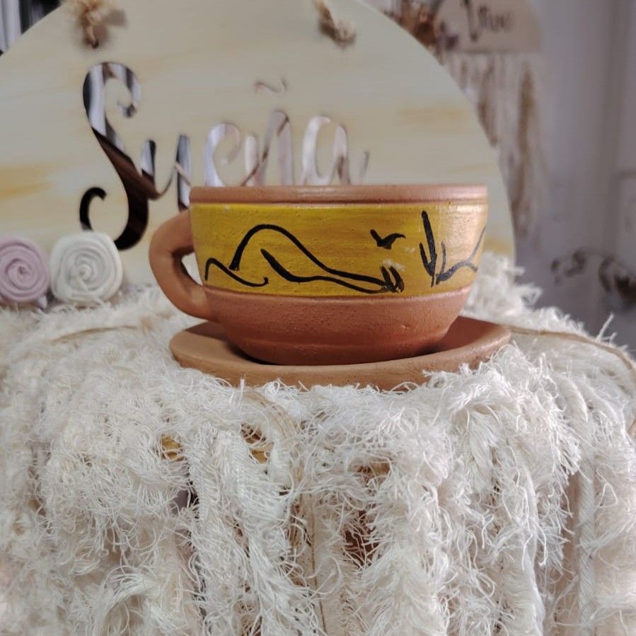 Taza de cerámica amarilla con platillo. La taza tiene una silueta negra. Sobre tela blanca afelpada.