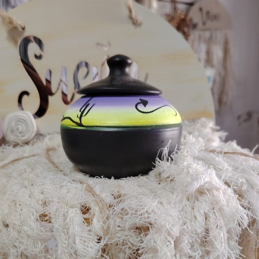Pequeño tarro negro con tapa morada y amarilla, decorado con diseños en negro, sobre tela texturizada de color blanco.