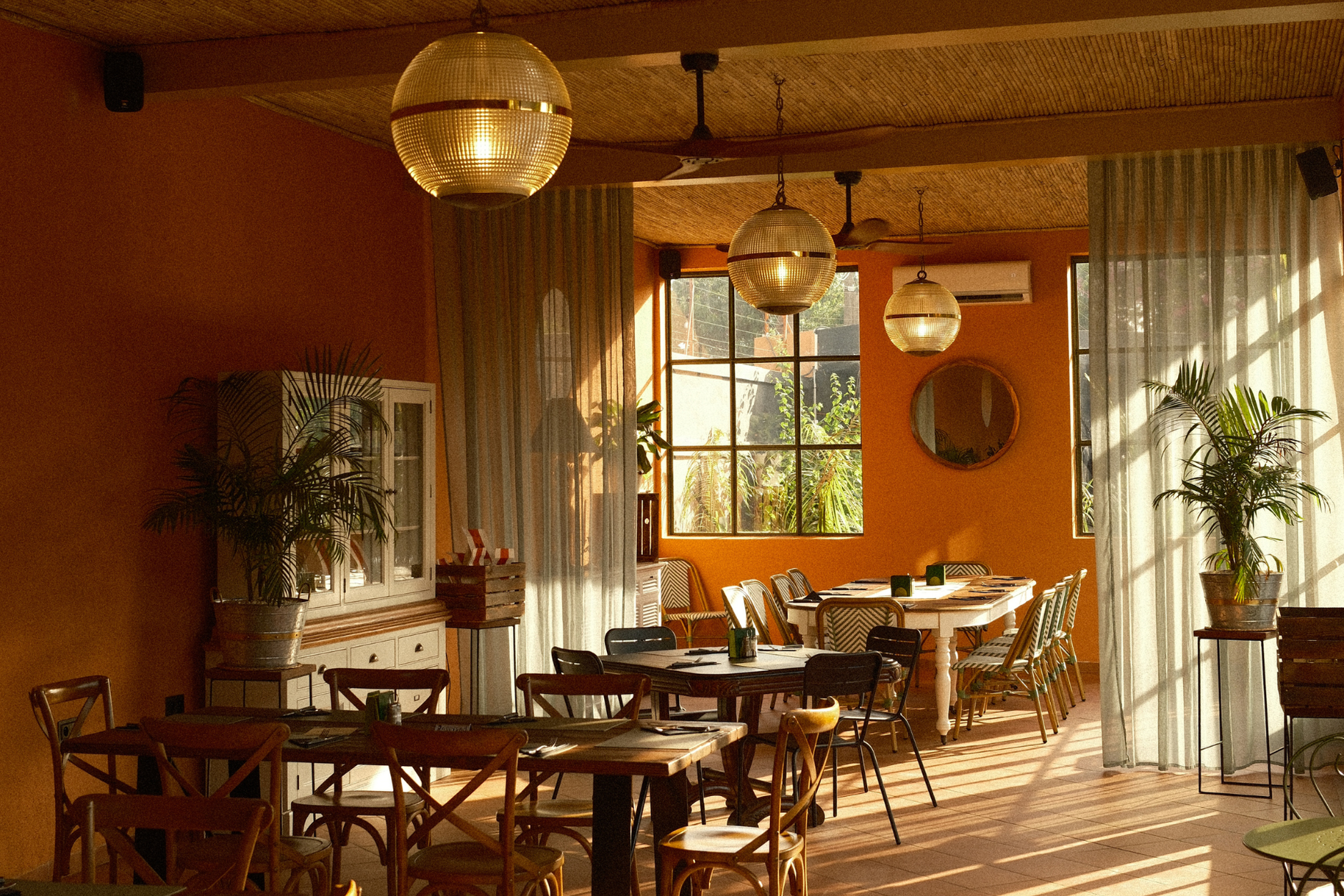 Intérieur d'un café avec tables et chaises, murs orange et grandes fenêtres. Des suspensions lumineuses et des plantes.