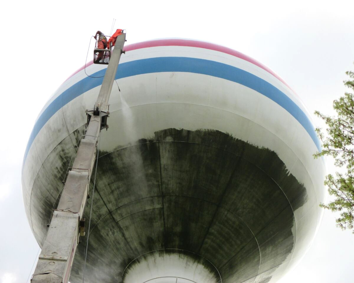 A person in a lift power washes a tall white water tower with red, white, and blue stripes.