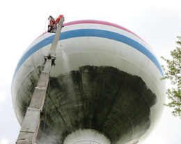 A person in a lift power washes a tall white water tower with red, white, and blue stripes.