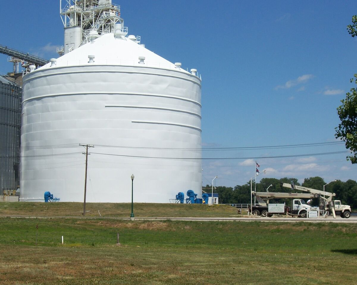 White grain silo with blue accents, power lines, truck on grassy field, sunny day.