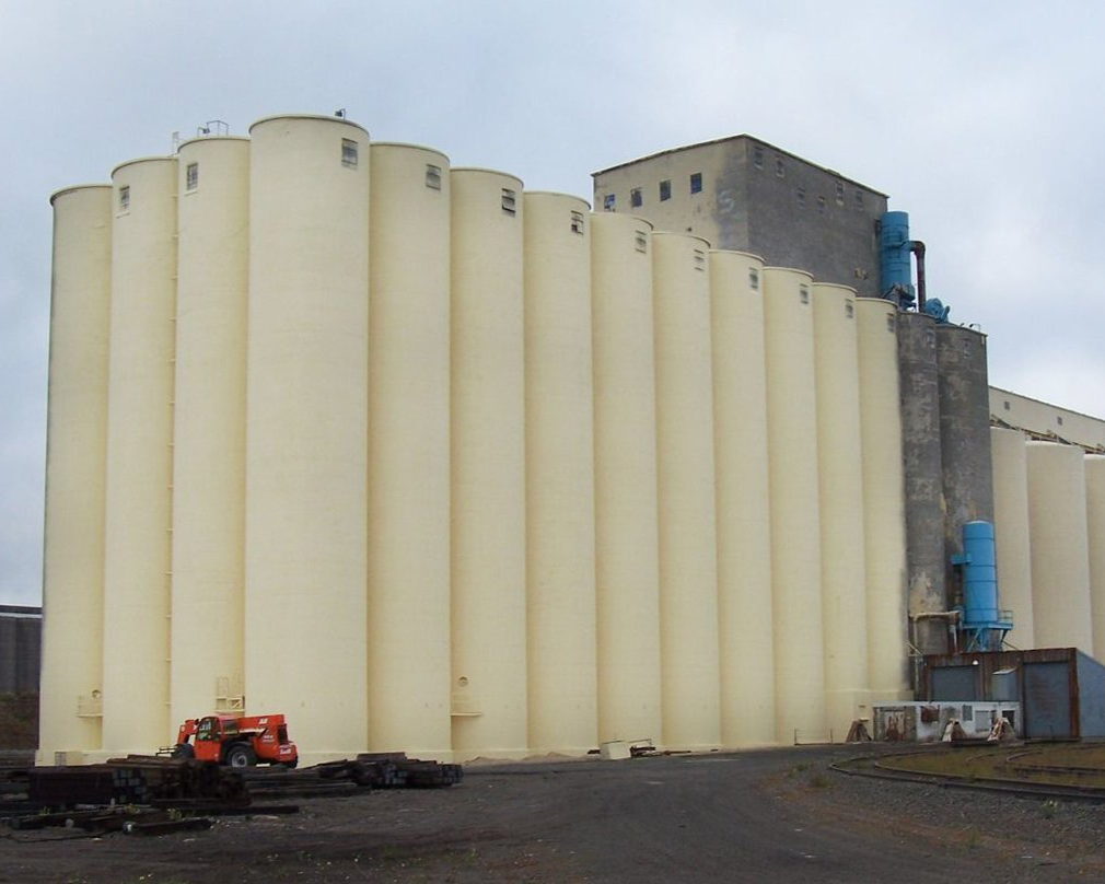 Large beige grain silos with a small orange forklift parked on a gravel surface.
