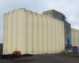 Large beige grain silos with a small orange forklift parked on a gravel surface.