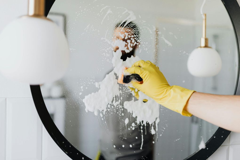 A person wearing yellow gloves is cleaning a mirror with a sponge.