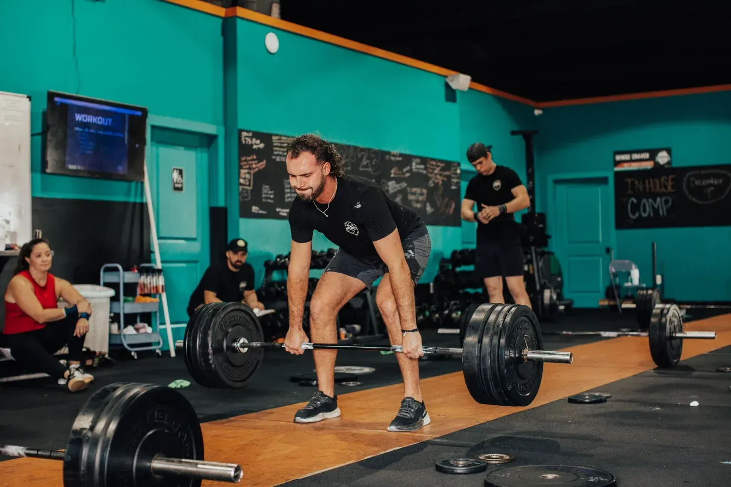 Man deadlifts barbell in a gym, others observing. Teal walls, black weights, determined expression.