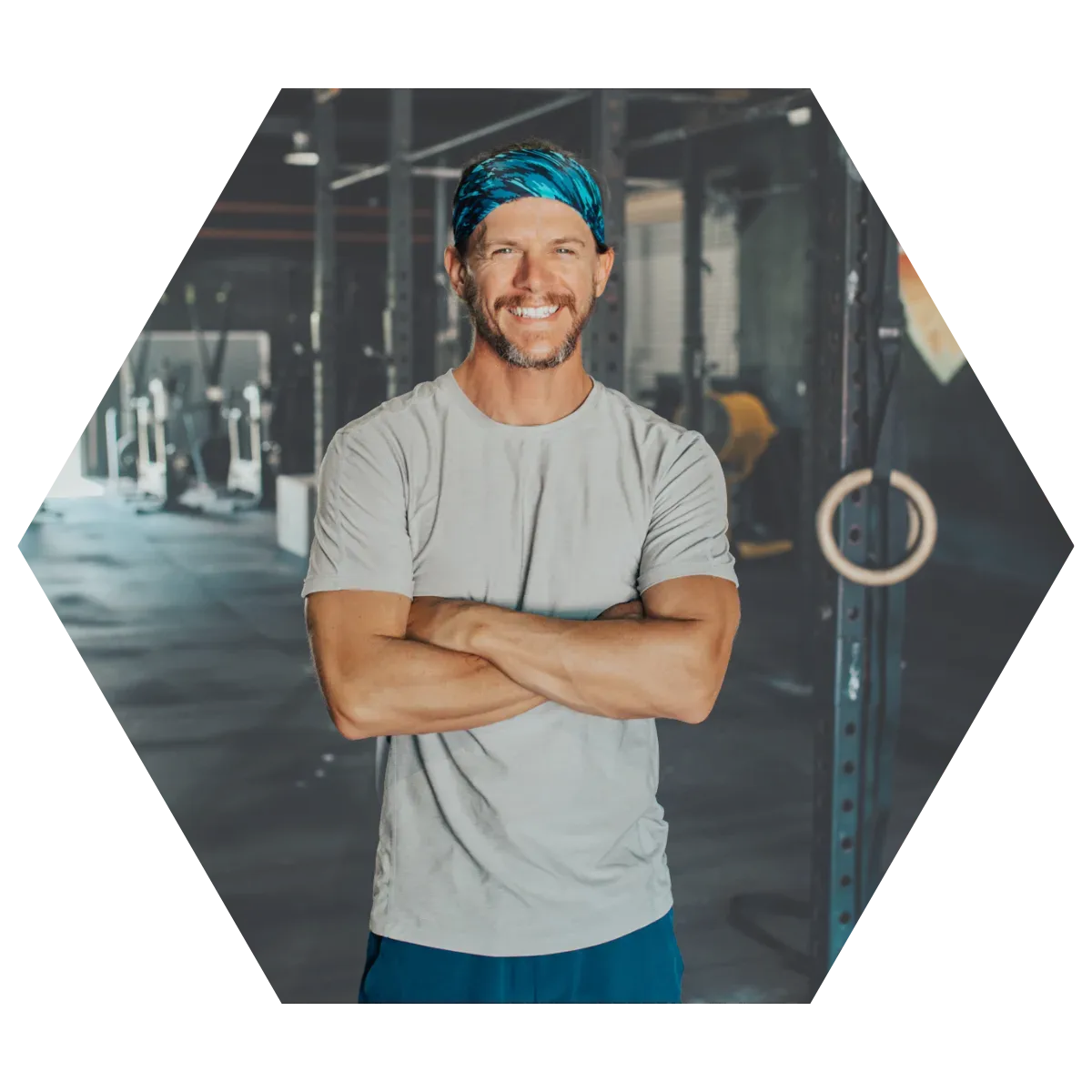 Man in a gym, arms crossed, wearing a blue bandana, light gray shirt, and dark blue shorts; smiling.
