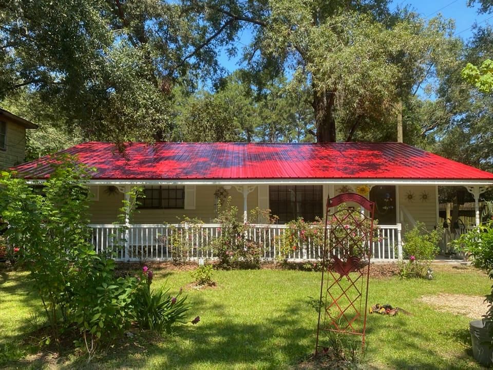 Cottage with red roof and white porch, surrounded by greenery, under a sunny sky.