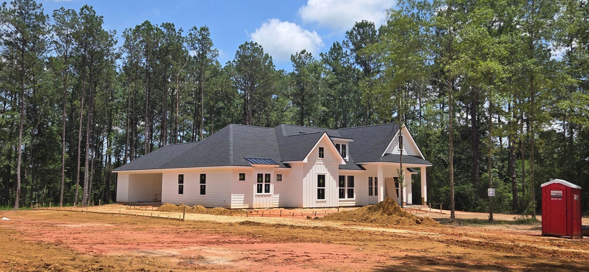 White house under construction with dark roof, dirt yard, and trees in the background. Red porta potty.