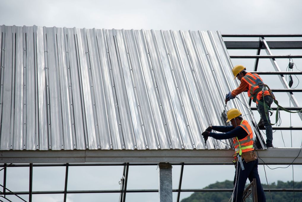 Two construction workers installing corrugated metal roofing on a building frame; cloudy sky.