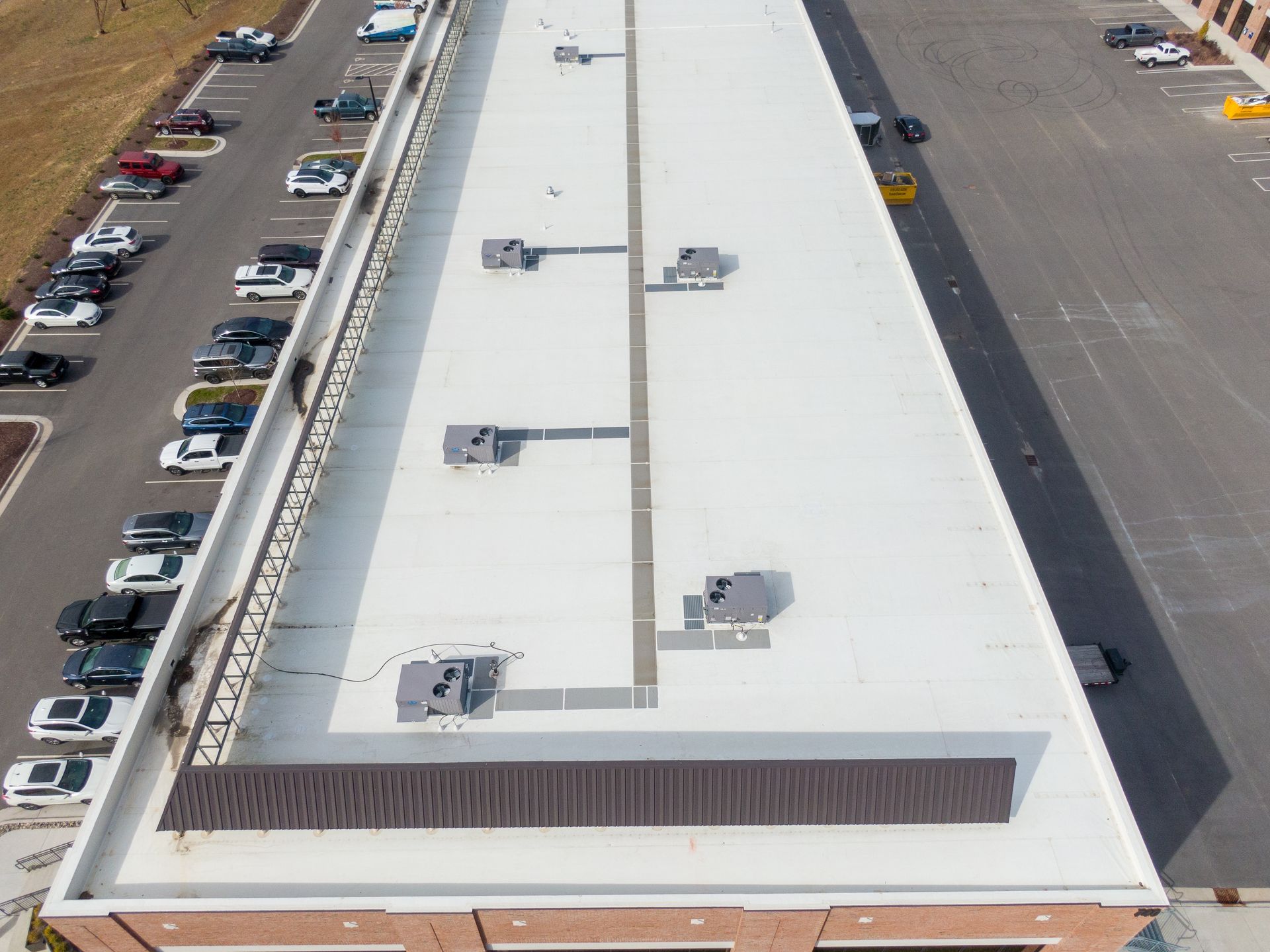 Aerial view of a building with a white flat roof, HVAC units, and a parking lot with cars.