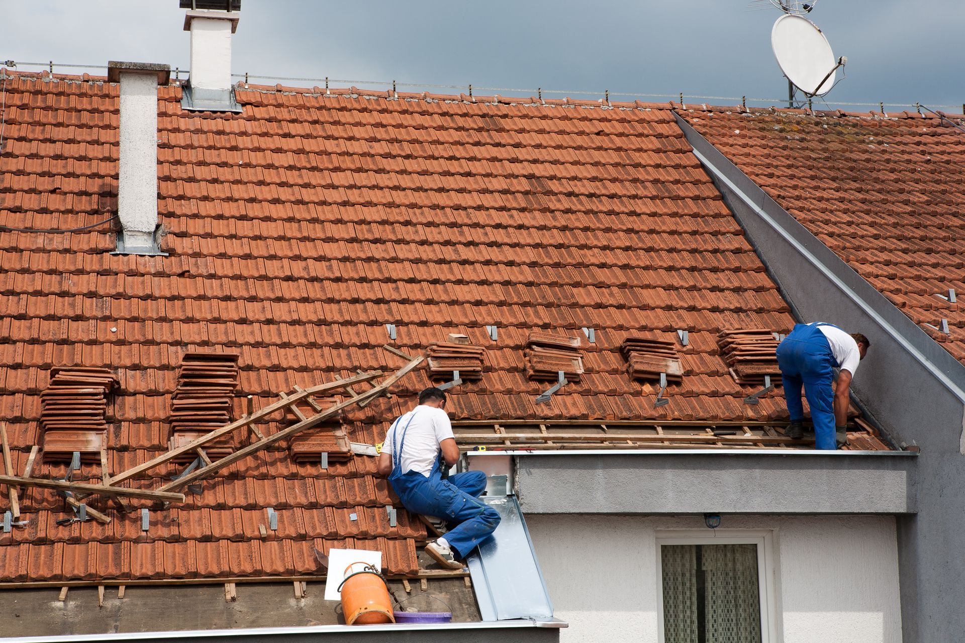 Two roofers in blue overalls repairing a terracotta tile roof on a building under a cloudy sky.