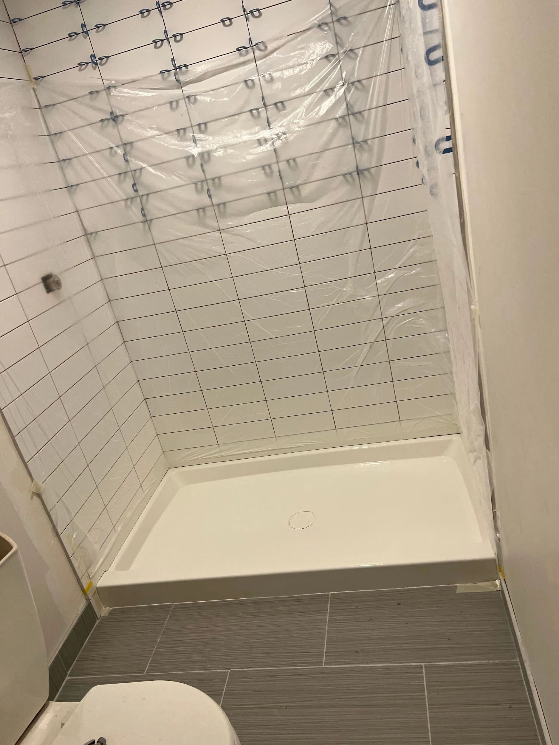 A walk-in shower with white tiled walls covered in plastic, a white shower base, and dark gray floor tiles.