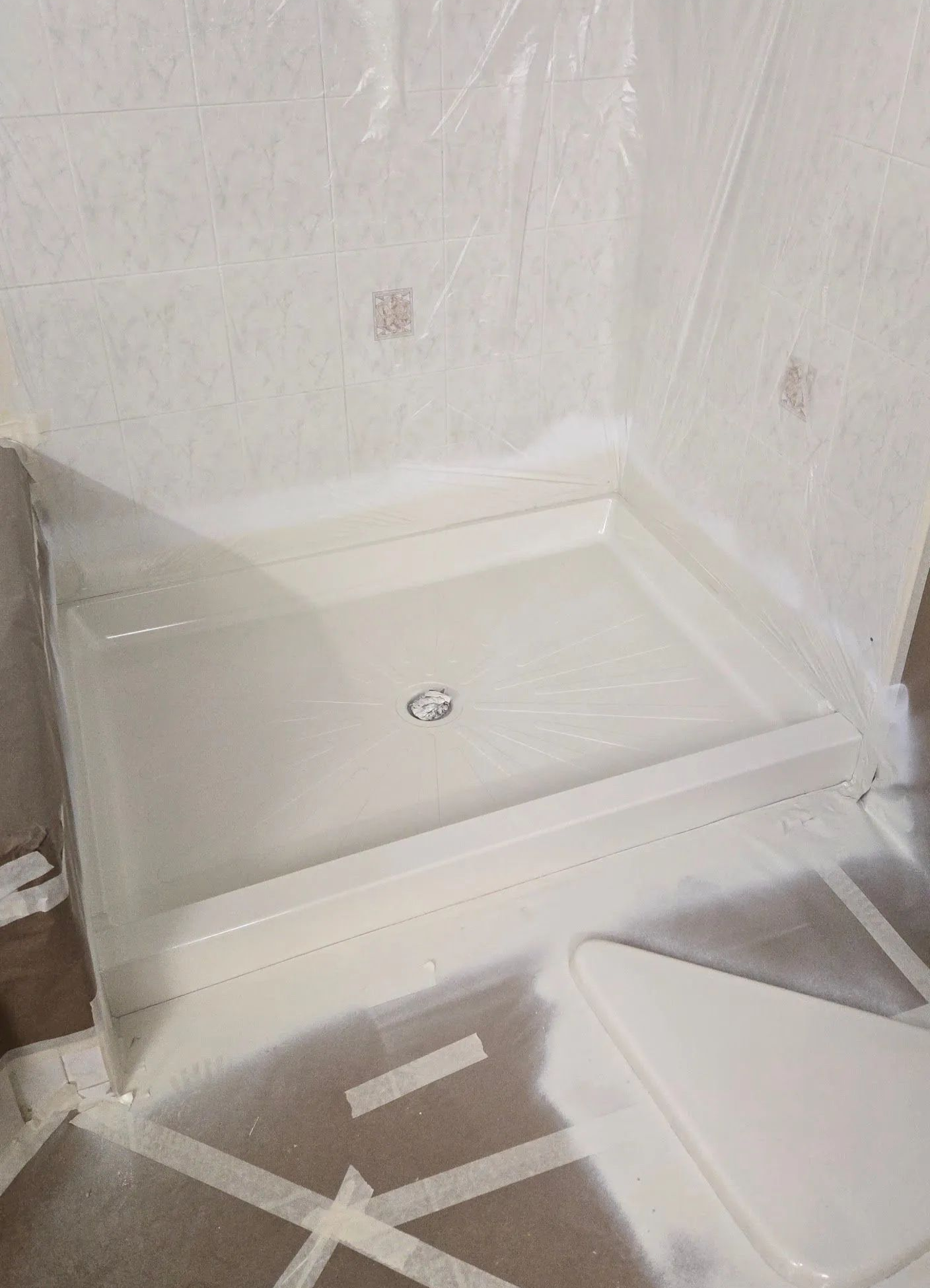 White corner shower base in a bathroom prepared for installation; protected with plastic.