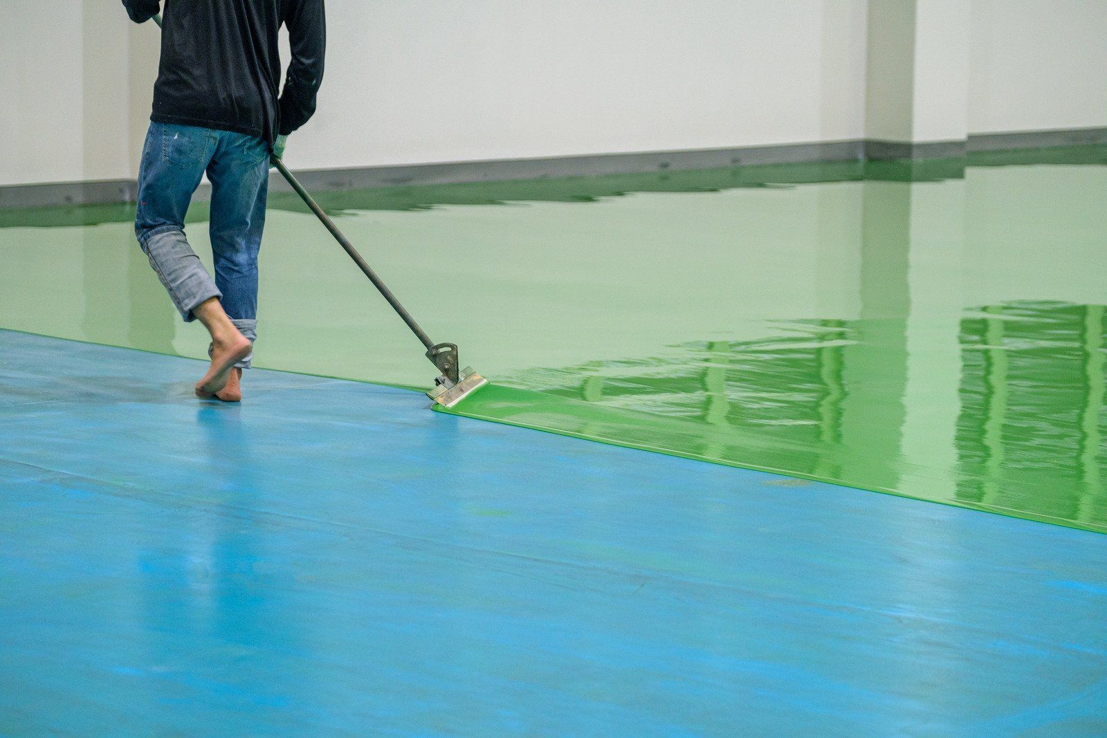Person applying green epoxy coating to a blue floor using a floor applicator in a warehouse.