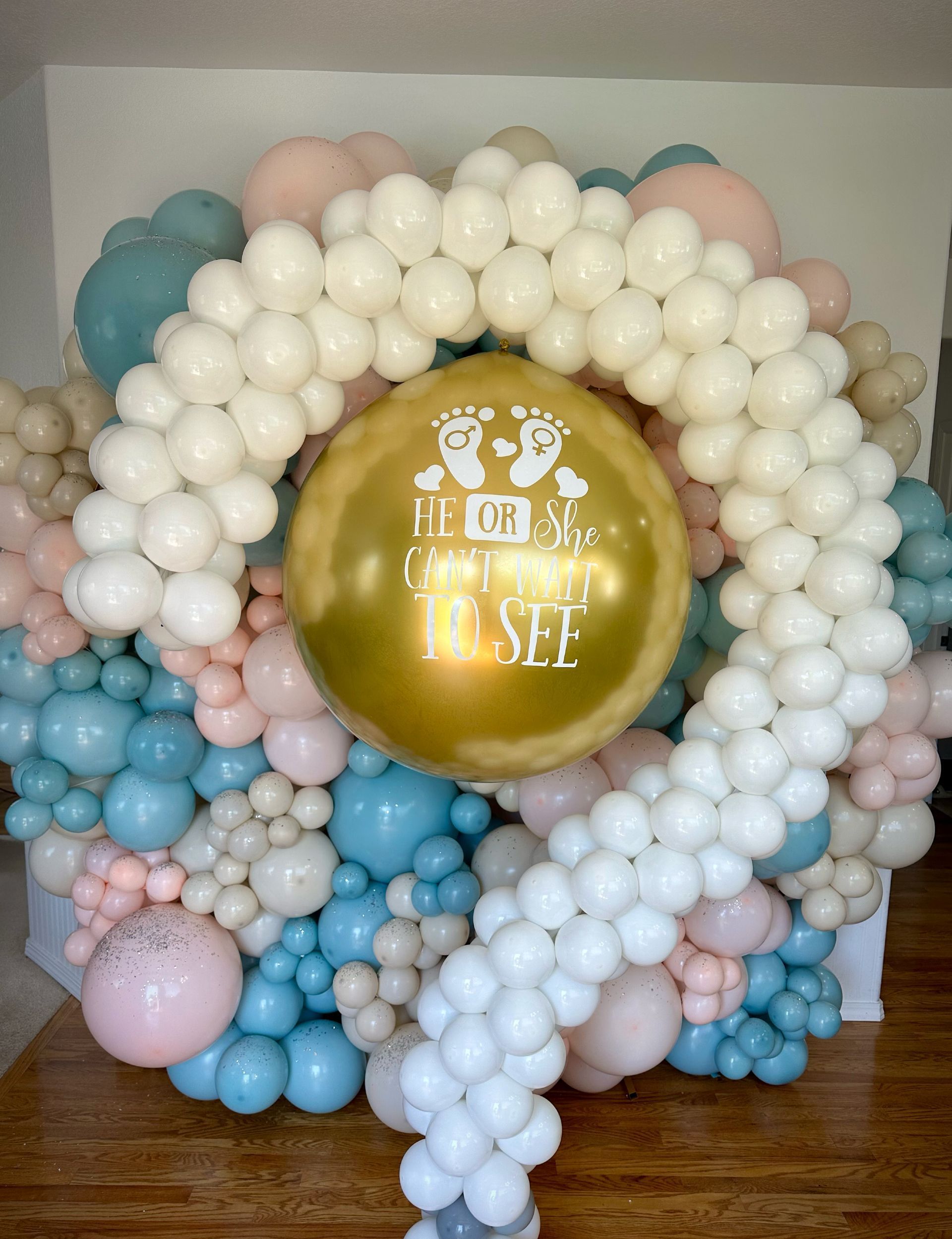 Gender reveal balloon display with white, pink, blue, and gold balloons. A gold balloon in the center reads 