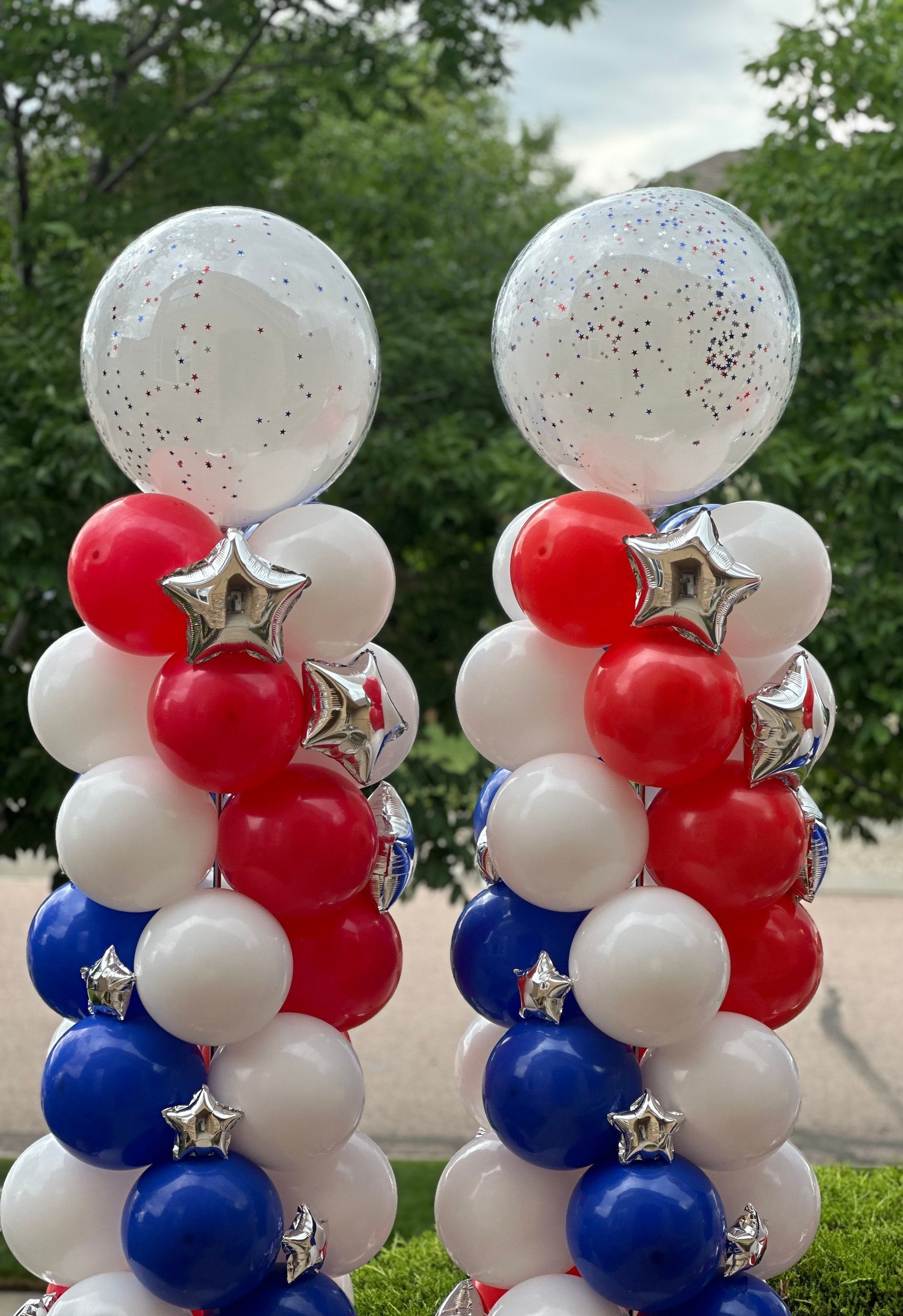 Two patriotic balloon columns; white, red, and blue balloons with silver star accents and large clear, glitter-filled top balloons.