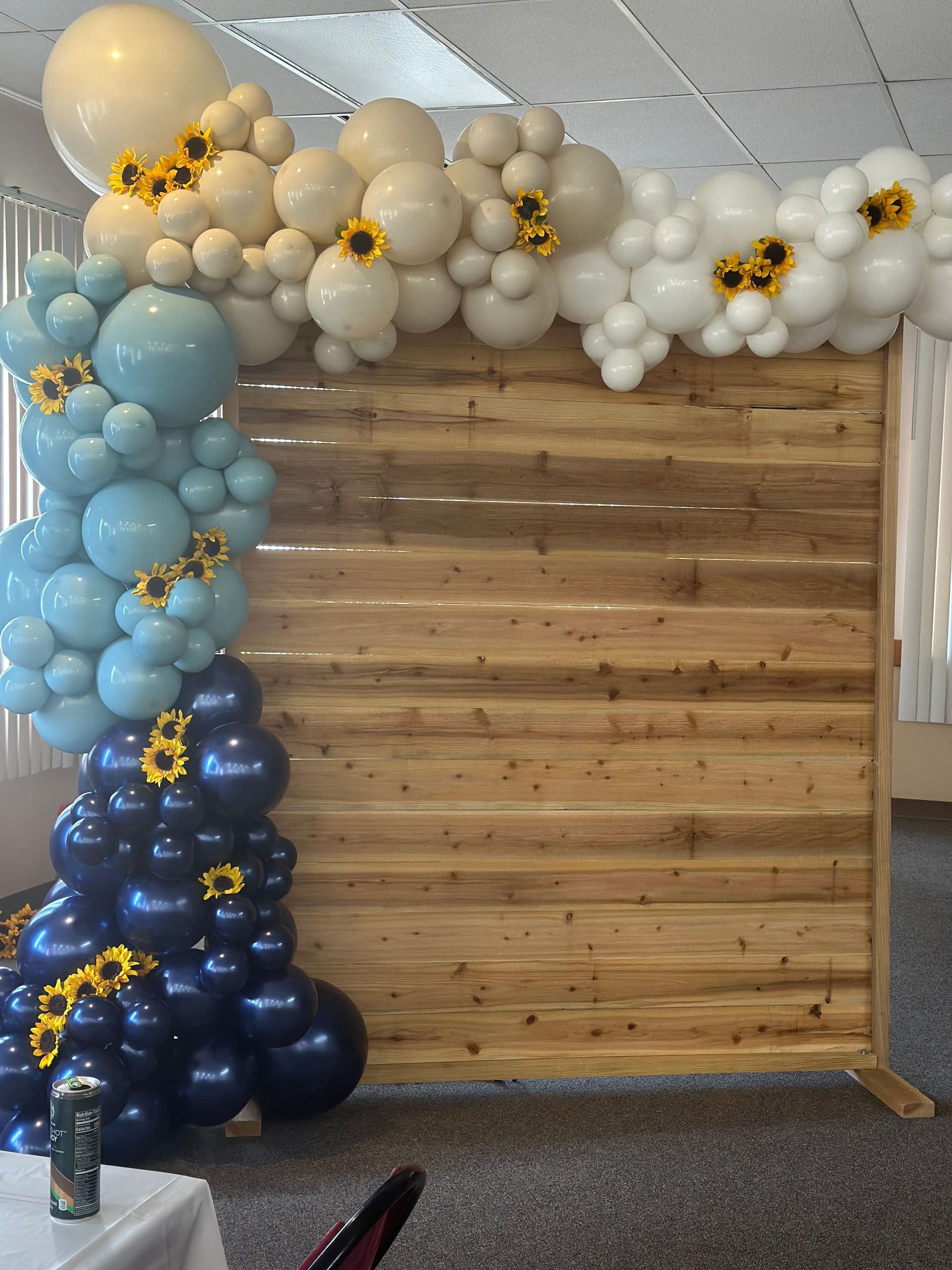 Balloon arch with sunflowers against a wooden backdrop. Blue, white, and tan balloons.
