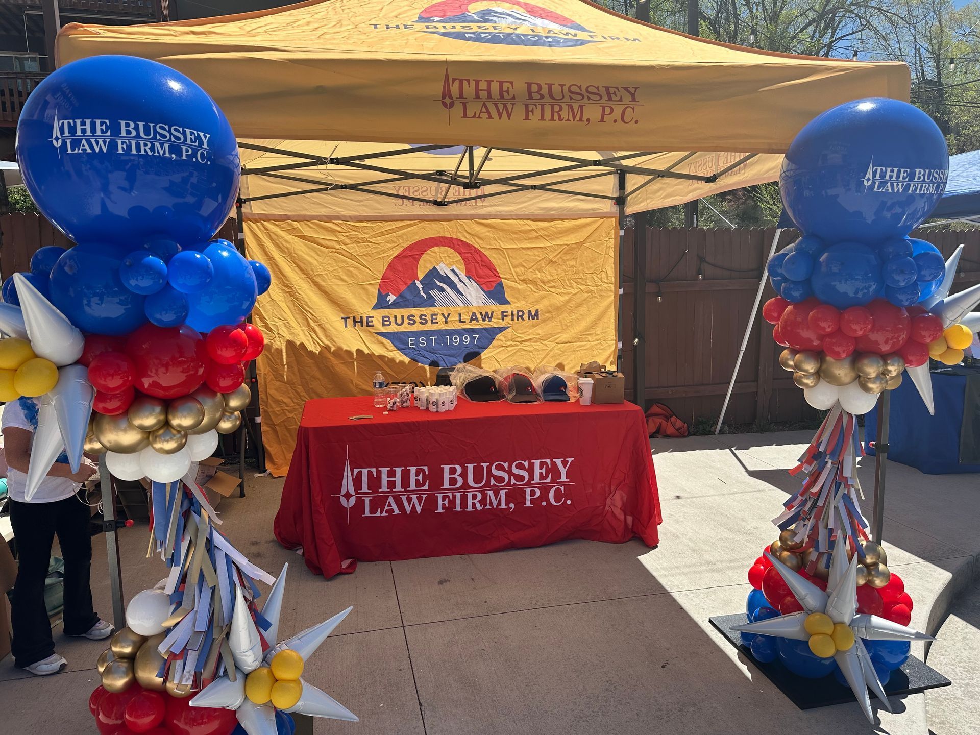 Law firm booth at an outdoor event, with red, white, and blue balloon decorations. Banner displays the firm's logo.