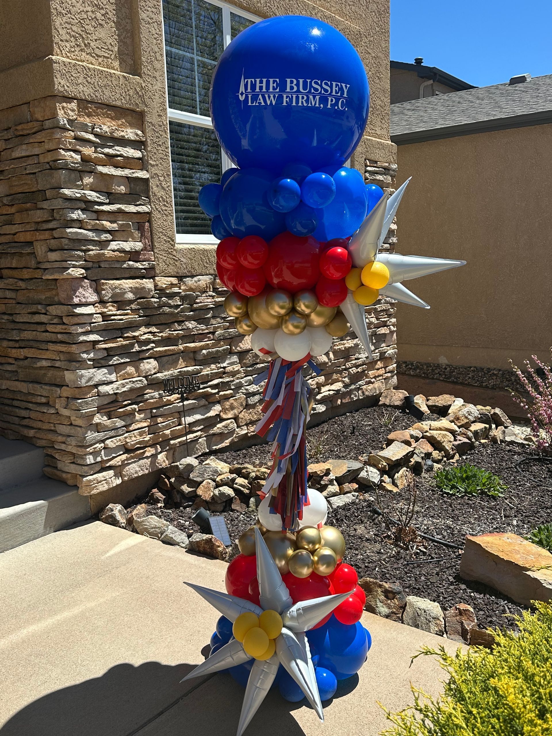 Balloon sculpture in red, white, blue, and gold, with star-shaped decorations, standing outside a building.