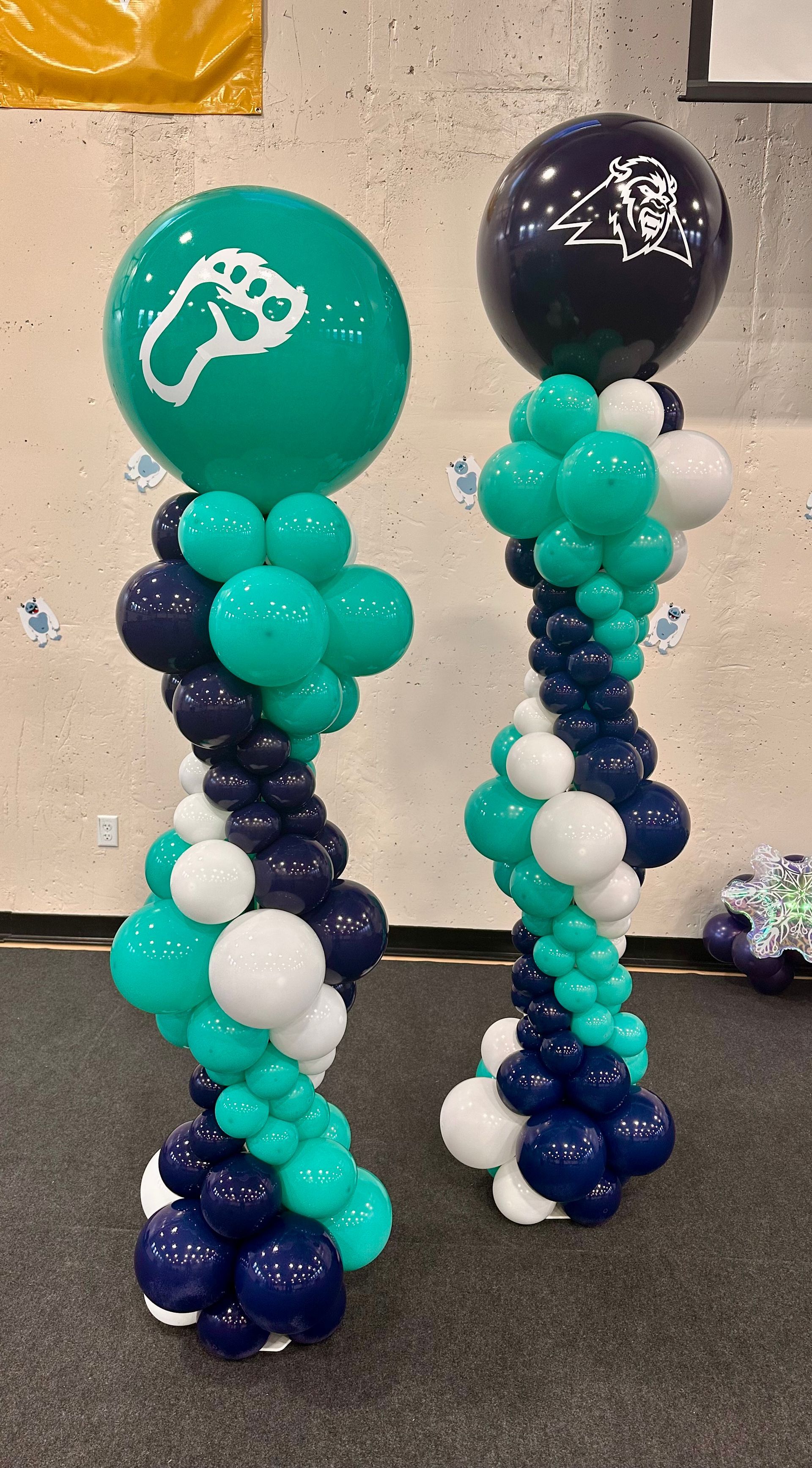 Two balloon columns: teal, navy, and white balloons in a spiral design, topped with round balloons; one has a footprint, the other an image.