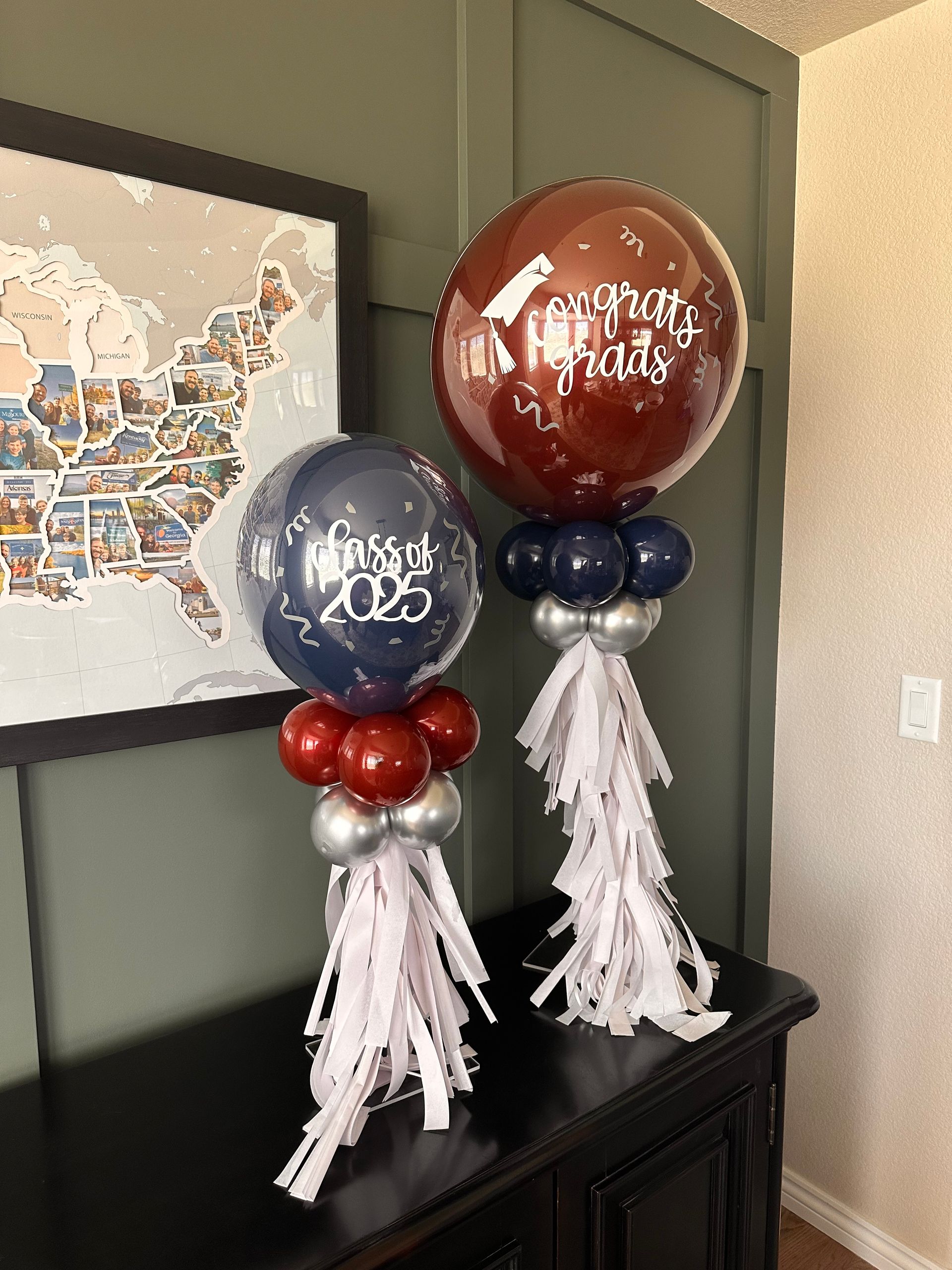 Two graduation-themed balloon arrangements with tassels, on a dark dresser, by a map.
