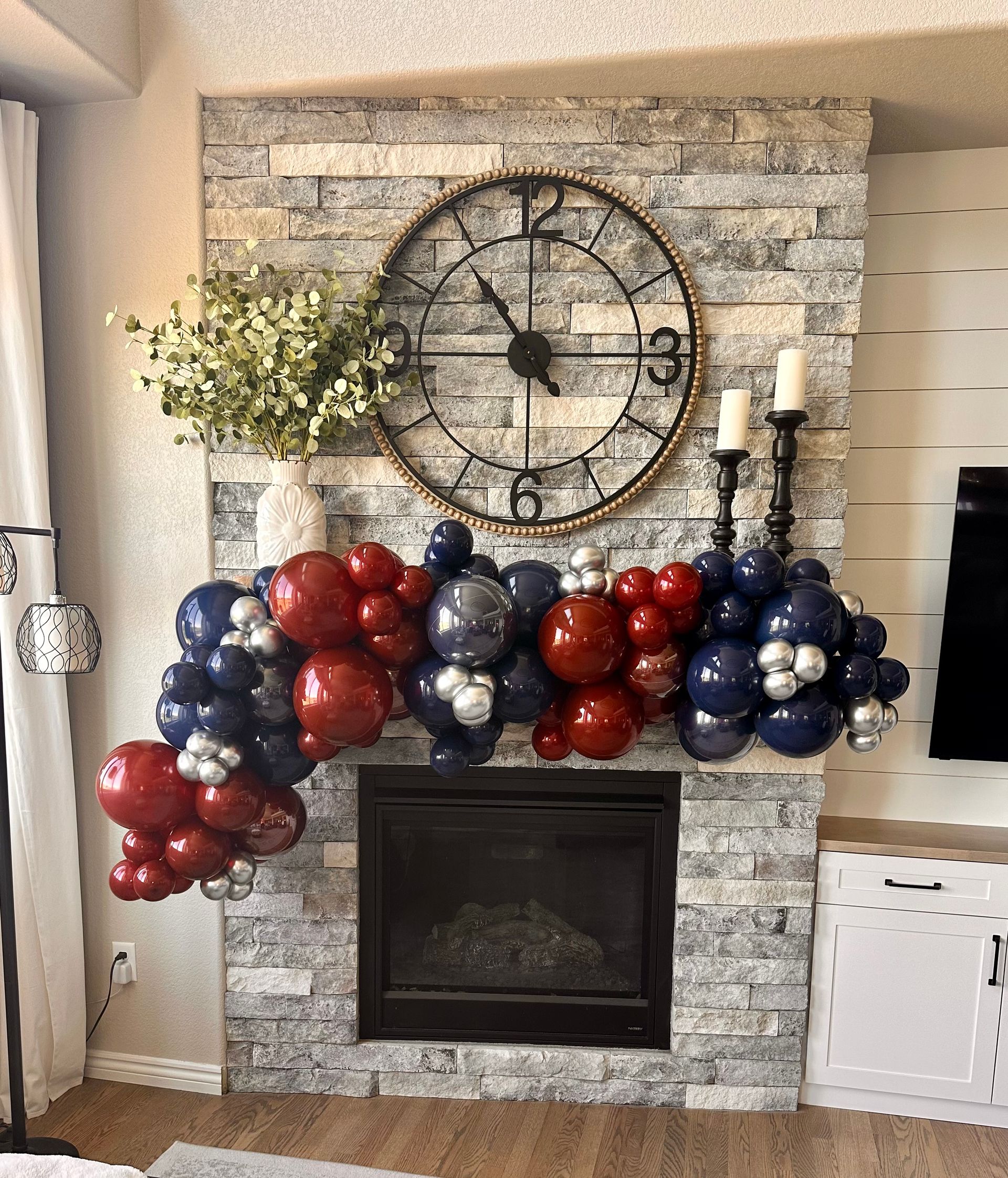Fireplace decorated with red, blue, and silver balloons, clock, greenery, and candles.