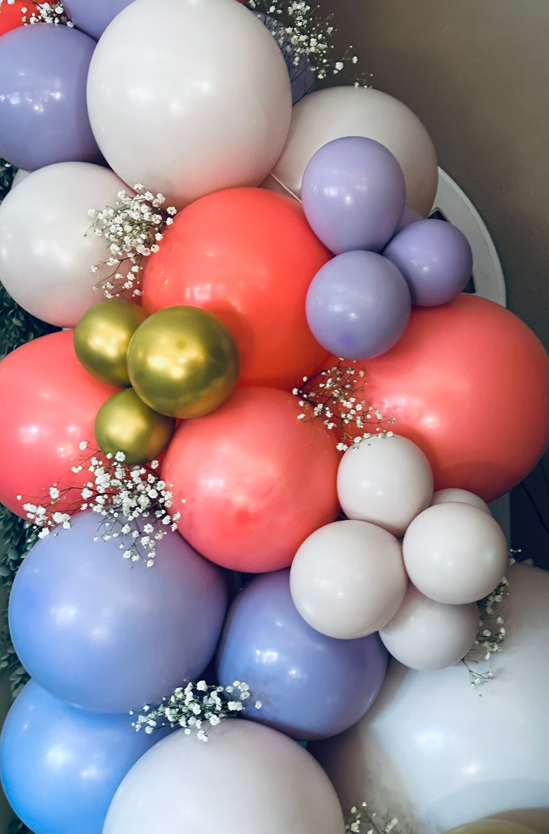 Balloon arrangement with coral, white, lavender, and gold balloons. Baby's breath accents.